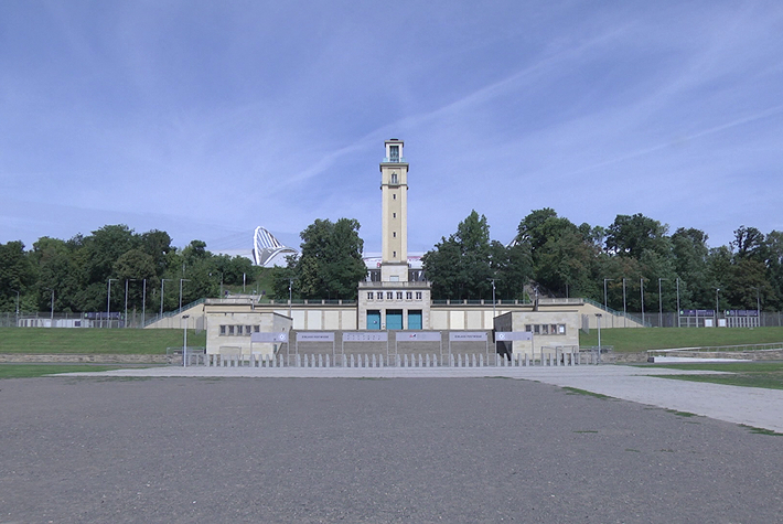Hier ist ein großer mit Kies versehener Bereich zu sehen, der von einem gepflasterten Gehweg umgeben ist, der wiederum von einer Grünfläche eingegrenzt wird. Mittig platziert ist ein Treppenaufgang und ein Turm zu erkennen.