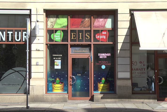 Hier ist mittig der verglaste Eingangsbereich des Eiscafés Grimms Eismärchen zusehen, dessen Logo auf der unteren linken und rechten Seite des Eingangs abgebildet ist. Außerdem ist über der Eingangstür in Großbuchstaben das Wort „Eis“ zu lesen.