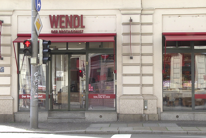 Hier ist die Glasfassade der Konditorei und Bäckerei Wendl zu erkennen, bei der sich links von der Eingangstür die Aufschrift „Der Brotagonist“ und rechts von der Eingangstür die Öffnungszeiten befinden. Über dem Eingang befindet sich der rote Schriftzug Wendl – Der Brotagonist.