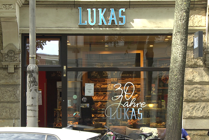 Abgebildet wird hier die Glasfassade der Bäckerei Lukas durch die man die Theke erkennen kann und auf der sich die Aufschrift „30 Jahre LUKAS“ befindet. Über dieser Glasfassade befindet sich zu dem der blaue in Großbuchstaben geschriebene Schriftzug „Lukas“.