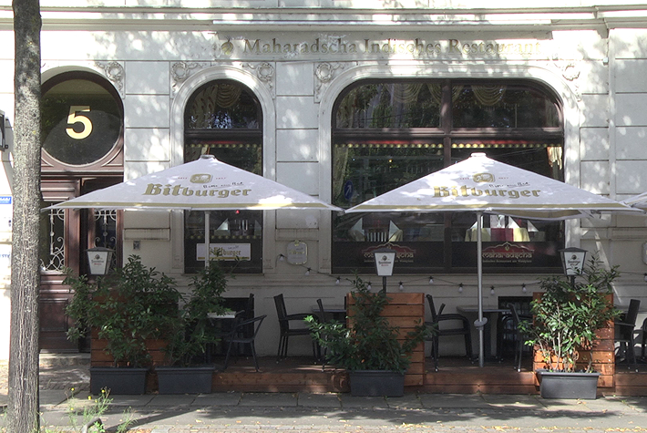 Hier ist ein Ausschnitt einer weißen Fassade mit dem goldenen Schriftzug „Maharadscha Indisches Restaurant“ zu erkennen. Davor befindet sich eine kleiner mit Pflanzen versehener Außensitzbereich des Restaurants.