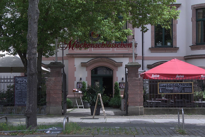 Hier ist ein Ausschnitt einer weiß-rötlichen Fassade zu erkennen, mit einem relativ großen davorliegenden Außensitzbereich. Über dem Eingangsbereich hängt der rote Schriftzug „Mückenschlösschen“.