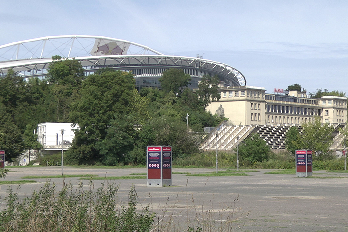 Hier ist eine seitliche Ansicht auf ein Teil eines Stadions zu sehen auf dem ein Plakat mit zwei roten Bullen zu erkennen ist. Vor dem Stadion ist ein Gebäude zu sehen, auf dem sich ein rot-blauer und in Großbuchstaben geschrieben Schriftzug „Red Bull Arena“ befindet.