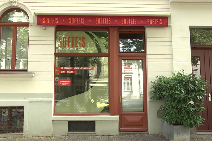 Hier ist der verglaste Eingangsbereich des Softeis Eugen Hendrich abgebildet, auf dem sich unter anderem der mit Großbuchstaben geschriebene Schriftzug „Softeis – Aus eigener Herstellung“ befindet. Über dem Eingangsbereich befindet sich ein Band aus rotem Stoff, auf dem mehrfach das Wort „Softeis“ zu lesen ist.