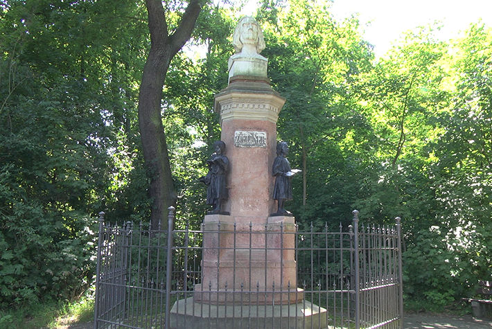 Hier ist das umzäunte Zöllnerdenkmal zu sehen, welches einen roten Sockel hat, auf dem schwarz aussehende Figuren mit Büchern in der Hand zu erkennen sind. In der Mitte ragt eine rötliche Säule heraus auf der ein weiße Büste platziert ist.